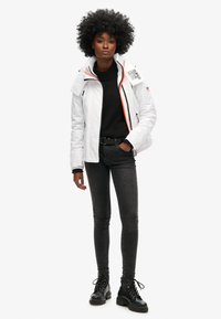 Biała kurtka izolacyjna z wysokim kołnierzem, czarnymi zamkami błyskawicznymi i wielokolorowym podszyciem. Połączona z czarnymi dżinsami i czarnymi butami sznurowanymi.