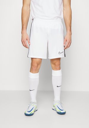 Homme portant un short de sport Nike blanc, des chaussettes hautes blanches avec logos Nike, et des chaussures de sport blanches avec des détails bleus et jaunes.
