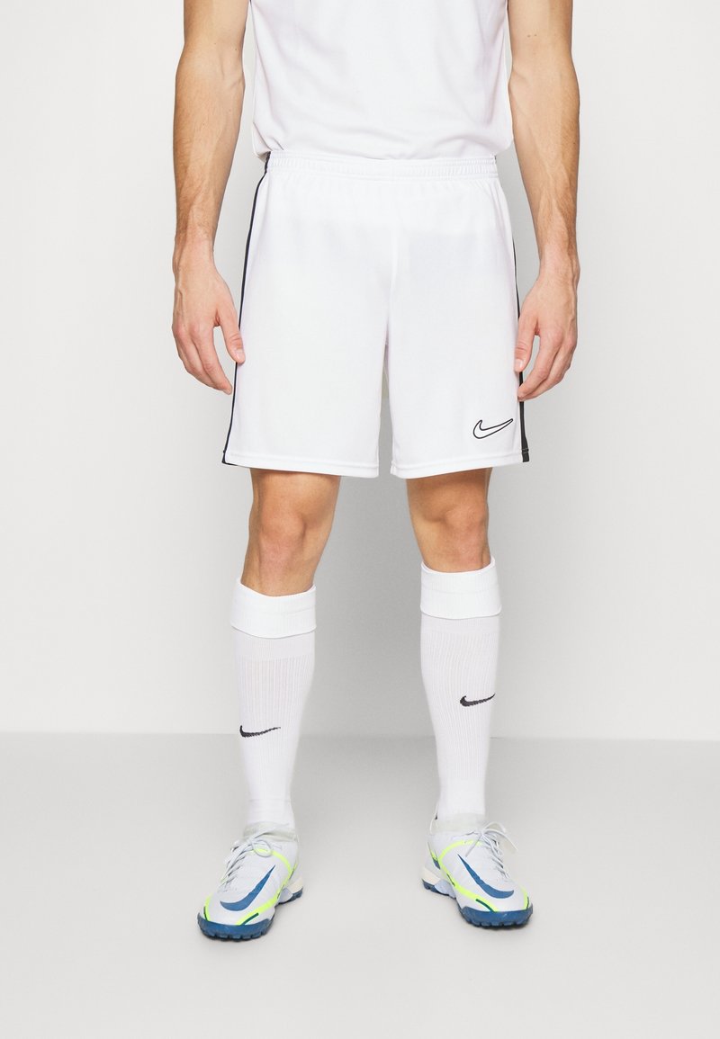 Homme portant un short de sport Nike blanc, des chaussettes hautes blanches avec logos Nike, et des chaussures de sport blanches avec des détails bleus et jaunes.