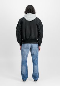 Black bomber jacket with a gray hoodie underneath, elastic cuffs, and waistband, paired with light blue jeans and black shoes.