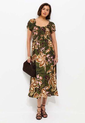 Kobieta w zielonej, kwiecistej sukience do połowy łydki z bufiastymi rękawami, trzymająca brązową plecioną torebkę i nosząca czarne sandały na paski.