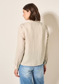 Camicia di lino beige con maniche lunghe e polsini abbottonati, caratterizzata da un orlo arrotondato e un tessuto testurizzato. Indossata con jeans azzurri chiari.
