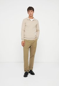 Beige gebreide trui met een kraag, gecombineerd met kaki broek. Model draagt zwarte schoenen. Effen achtergrond.