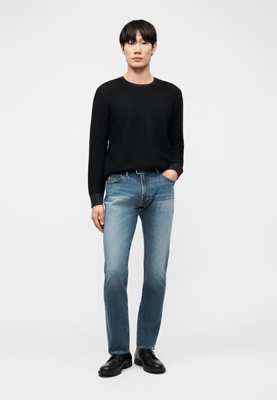 Άνδρας που στέκεται μπροστά σε απλό φόντο, φοράει μαύρο πλεκτό πουλόβερ με ριμπ ύφασμα, μπλε τζιν και μαύρα παπούτσια, με το ένα χέρι στην τσέπη.