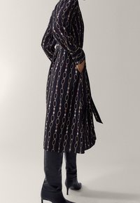 Musta midi-mekko, jossa on ketjumalli, pitkät hihat, kapea vyötärö, sivutaskut, yhdistetty polvipitkiin mustiin saappaisiin. Sametintuntu.