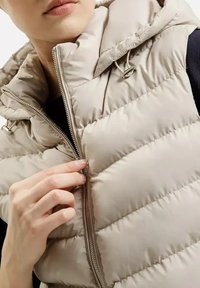 Beżowa kamizelka puchowa z kapturem. Posiada zamek błyskawiczny, przeszywane wzory oraz sznurek przy dekolcie. Miękka, pikowana tekstura.