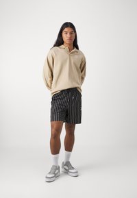 Beige Langarm-Polo mit Kragen und weißem Logo, kombiniert mit schwarz-weiß gestreiften Shorts. Das Model trägt graue Sneakers und weiße Socken.