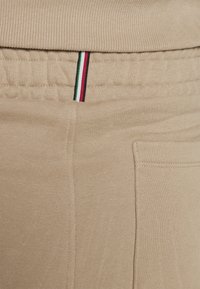 Beige Sweatpants mit geripptem Bund, Seitentasche und einem dekorativen gestreiften Akzent in Grün, Rot und Schwarz an der Oberseite.
