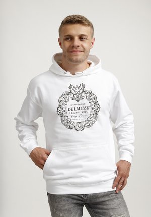 Młody mężczyzna w białej bluzie z kapturem z czarnym dekoracyjnym logo „Champagne De Lalisse Grand Cru”, stoi z rękami w kieszeniach, lekko się uśmiechając.