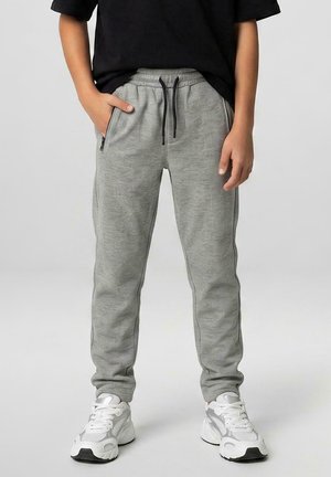 Kind draagt grijze joggingbroek met patroon en zwarte trekkoorden en witte sneakers, staat met één hand in de zak.