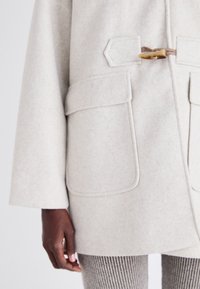 Manteau en laine gris clair à finition texturée, comportant de grandes poches, une fermeture à ceinture et un léger éclat. Les accents incluent un bouton en bois.