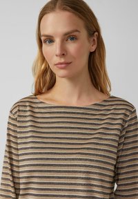 Frau mit schulterlangem, blonden Haar, die ein beige und braun gestreiftes Oberteil trägt und leicht zur Seite schaut, vor einem schlichten Hintergrund.