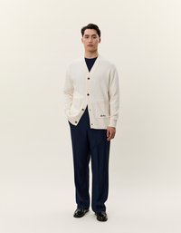 Cardigan grosso in crema con scollo a V, bottoni in legno, due tasche frontali e brandizzazione ricamata, abbinato a pantaloni blu navy e scarpe nere.