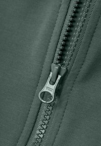 Tejido verde con una cremallera YKK, con un acabado texturizado. El tirador de la cremallera es metálico con un logo grabado. Superficie lisa alrededor.