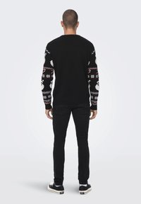 Svart genser med mønstrede ermer som har hvite og røde design; kombinert med svarte jeans og svarte joggesko. Bakre utsikt.