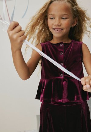 Burgunderrotes Samt-Peplum-Top mit Knöpfen, kombiniert mit einem passenden Rock. Das Kind hält ein weißes Band, die Haare sind wellig und schulterlang.
