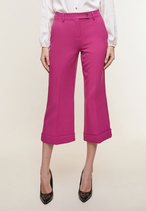 Vrouw in felroze wijde cropped broek met opgerolde zomen, gecombineerd met zwarte pumps met puntige neus en een wit blouse met knopen.