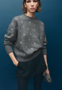 Szary, dziergany sweter z kwiatowymi ozdobami z koralików, okrągły dekolt, luźny krój, zestawiony z ciemnymi szortami i fakturowaną kopertówką.