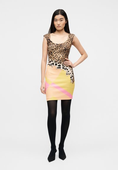 Γυναίκα με μακριά σκούρα μαλλιά, φορούν μια αμάνικη μπλούζα με ζωικό μοτίβο και μια πολύχρωμη μίνι φούστα με σχέδια, μαύρο καλσόν και μαύρες γόβες.