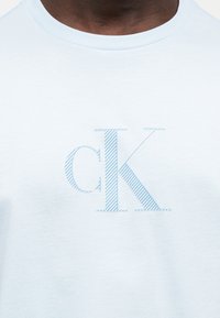 Άτομο με σκούρο δέρμα φοράει λευκή μπλούζα με στρογγυλή λαιμόκοψη και ελαφρώς γαλάζιο, διαγώνια ριγέ λογότυπο "cK" στο κέντρο του στήθους.