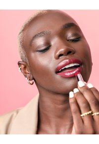 Red lip gloss being applied to full lips. The individual has a smooth, dark brown complexion and short, blonde hair with a textured finish.