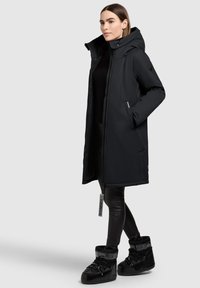 Czarna parka z kapturem, długimi rękawami i przednimi kieszeniami. W połączeniu z czarnymi legginsami z imitacji skóry oraz czarnymi botkami z futrzanym obszyciem.