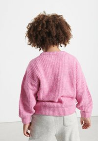 Ružový pletený sveter s textúrovaným vzorom, vo voľnom strihu a s rebrovanými manžetami, skombinovaný so sivými teplákmi. Pohľad zozadu.