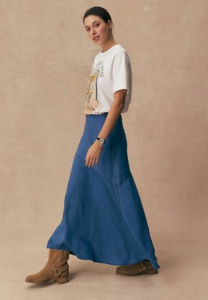 Femeie care merge pe jos, purtând un tricou alb cu imprimeu, o fustă lungă albastră, ghete maro până la gleznă și un ceas de mână, pe un fundal simplu bej.