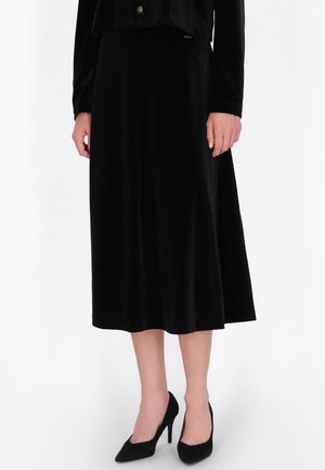 Una modelo lleva una blusa negra de manga larga, una falda hasta media pantorrilla y zapatos de tacón alto y punta afilada, sobre un fondo blanco de estudio.