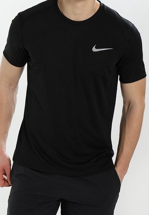 Homme portant un t-shirt de sport Nike noir à manches courtes avec un logo swoosh gris sur la poitrine, debout avec une main dans la poche.