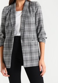 Šedý kostkovaný blazer s přiléhavým střihem, ohrnutými rukávy a jednou přední kapsou, spárovaný s bílou košilí a černými kalhotami.