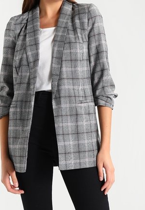 Grijze geruite blazer met een getailleerde pasvorm, opgerolde mouwen en een enkele voorzak, gecombineerd met een wit shirt en zwarte broek.
