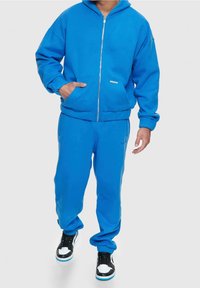 Blaue_zip-up-Hoodie_mit_Taschen,_kombiniert_mit_passenden_Sweatpants._Besitzt_eine_einfarbige_Ausführung,_weichen_Stoff_und_einen_lässig_geschnittenen_Schnitt._Schwarz-weiße_Sneaker.