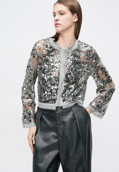 Ασημί μπουφάν cropped με παγιέτες, διαφανές ύφασμα, ξεφτισμένες άκρες και στρογγυλή λαιμόκοψη. Το μοντέλο το φοράει πάνω από σκούρες δερμάτινες ψηλόμεσες παντελόνες.