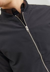 Czarna kurtka bomberka o satynowym wykończeniu, z prążkowanym kołnierzem i srebrnym metalowym zamkiem błyskawicznym o delikatnym połysku. Pod spodem znajduje się ciemna koszula.