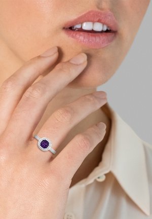Silberring mit einem kissenförmigen lila Edelstein, umgeben von kleinen weißen Steinen, getragen an einer Hand mit neutralem Nagellack, vor einem hellen Hintergrund.