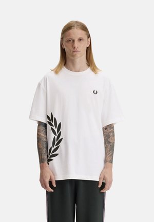 Joven con cabello largo y rubio que lleva una camiseta blanca de gran tamaño con estampado negro de laurel en el lateral y pantalones negros con franjas en los laterales.