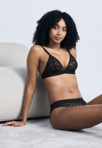 Femme aux cheveux bouclés assise sur un tapis clair, portant un soutien-gorge en dentelle noire transparente et un shorty assorti, appuyée contre un canapé blanc texturé.