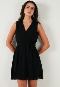 Zwart wikkeljurk met een V-halslijn, mouwloos ontwerp en een aangesloten taille. Voorzien van een strikceintuur en een vloeiende rok die tot halverwege de dij reikt.