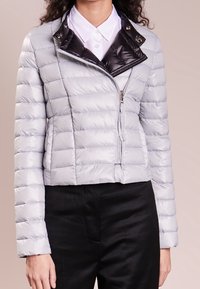 Femme portant une veste matelassée gris clair avec une fermeture éclair asymétrique et un col noir brillant par-dessus une chemise blanche à col.