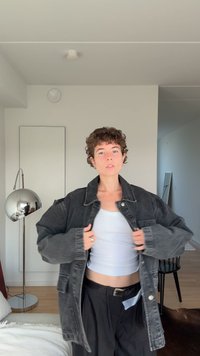 Oseba s kratkimi kodrastimi lasmi, oblečena v belo kratko majico in prevelik črni denim jakni, stoji v sodobnem minimalističnem prostoru.