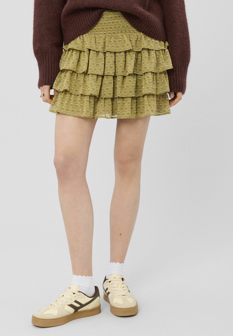 Mini-jupe courte à volants à motifs floraux verts, associée à des baskets beiges et des chaussettes blanches, portée avec un pull bordeaux.