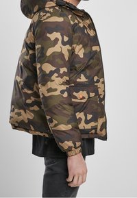 Kurtka puchowa w kamuflażu w odcieniach zieleni, brązu i czerni, z kapturem, zamek błyskawiczny, elastyczne mankiety oraz boczna kieszeń z pomarańczowym akcentem.