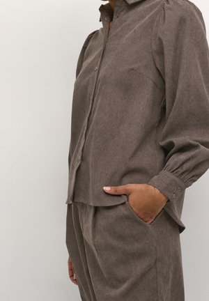 Woman wearing loose brown button-up shirt and matching pants with hand in pocket against plain light background.