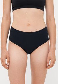 Woman wearing black high-waist underwear and a black crop top, standing with arms relaxed against a plain white background.