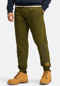 Olivgrüne Jogginghosen verfügen über einen Kordelzugbund, Seitentaschen und den Schriftzug "Timberland" mit einem Logo nahe dem Knöchel, kombiniert mit beigen Stiefeln.
