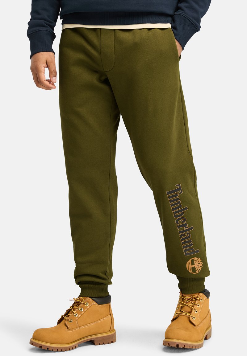 Olivgrüne Jogginghosen verfügen über einen Kordelzugbund, Seitentaschen und den Schriftzug "Timberland" mit einem Logo nahe dem Knöchel, kombiniert mit beigen Stiefeln.