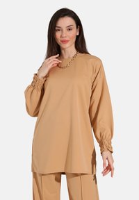 Langærmet skjorte lavet af glat beige stof, med elastiske manchetter og en afslappet pasform, suppleret med en kraftig kædehalssmykke.