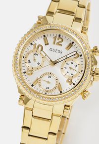 Gold-tone Guess wristwatch with a metallic link band, silver dial, three subdials, and crystal accents on the bezel and markers.