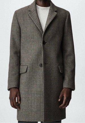 Mann trägt einen braunen und grauen Mantel in Knitterhauer-Muster mittlerer Länge mit Revers, zwei vorderen Patentaschen und Knopfleiste über einem hellen Pullover.